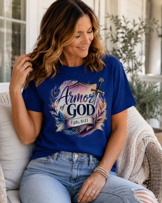 A woman sitting on a porch wearing a royal blue graphic t-shirt that reads 'Armor of GOD Eph. 6:11' with a shield and sword design, paired with light-wash jeans.