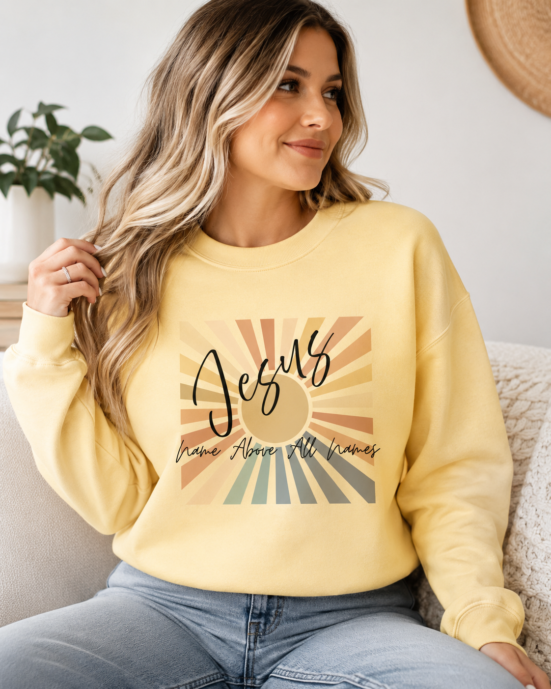 A woman wearing a pale yellow crewneck sweatshirt with a retro-style graphic that reads 'Jesus Have Mercy, All Knaves' over a sunburst design, paired with light blue jeans in a cozy indoor setting.