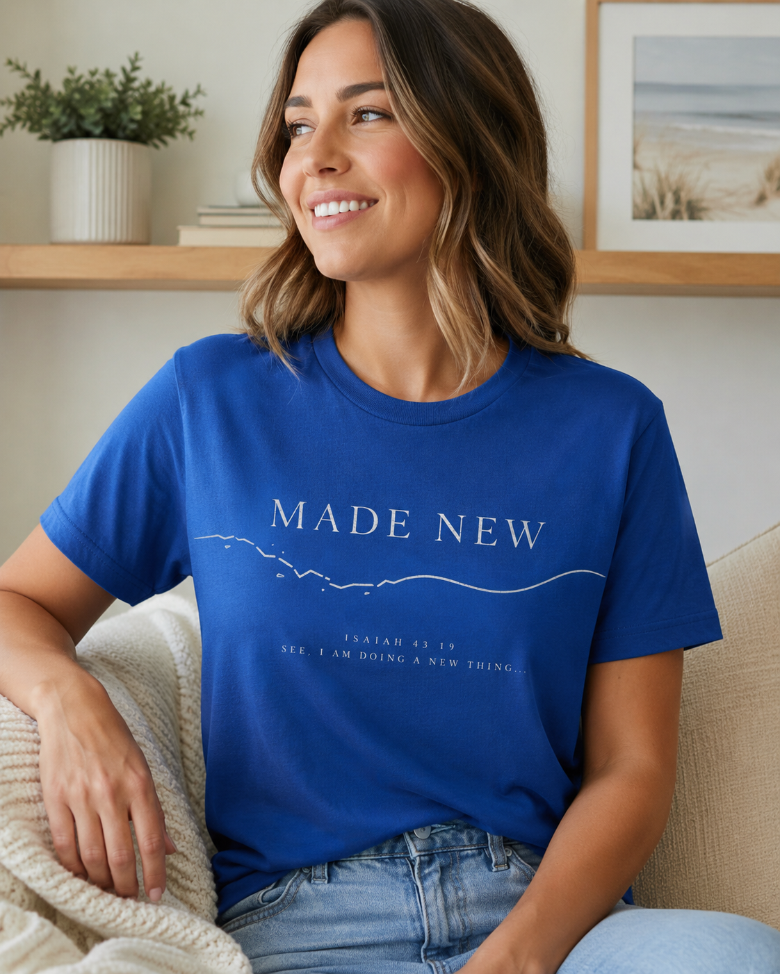 A woman sitting on a beige couch wearing a royal blue graphic t-shirt with the text 'MADE NEW' and 'JASIAN 4:18 SEE I AM DOING A NEW THING.' in white, paired with light-wash jeans.