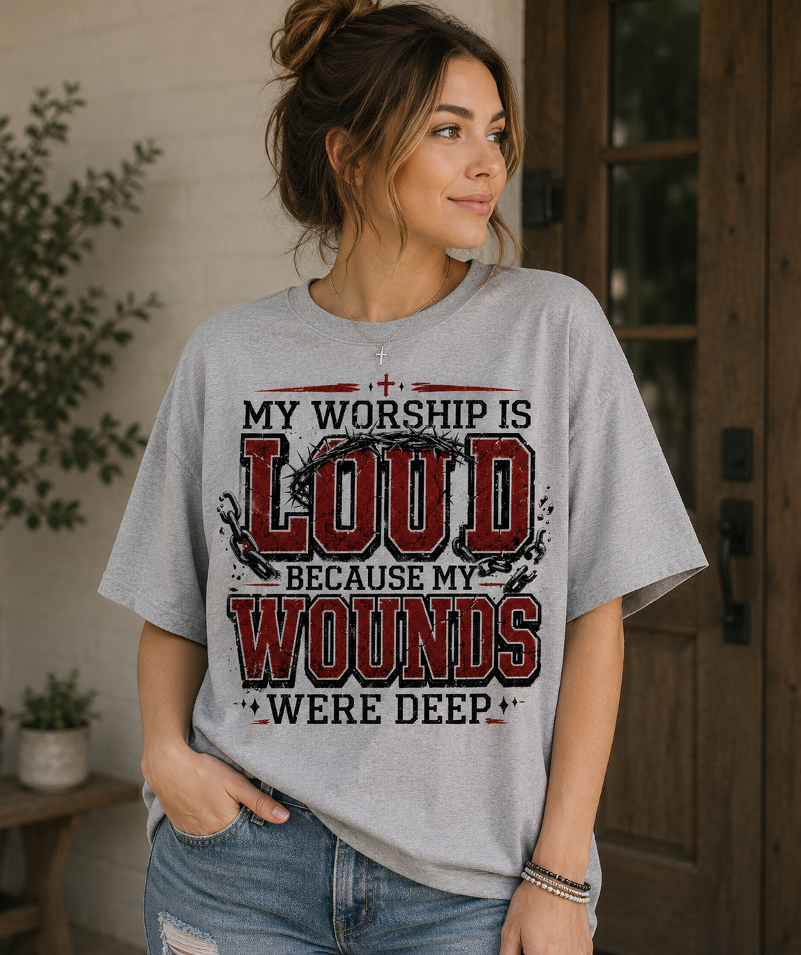 Woman wearing a gray My Worship Is Loud Because My Wounds Were Deep Christian T-shirt with distressed red and black lettering, crown of thorns, broken chains, and cross details.
