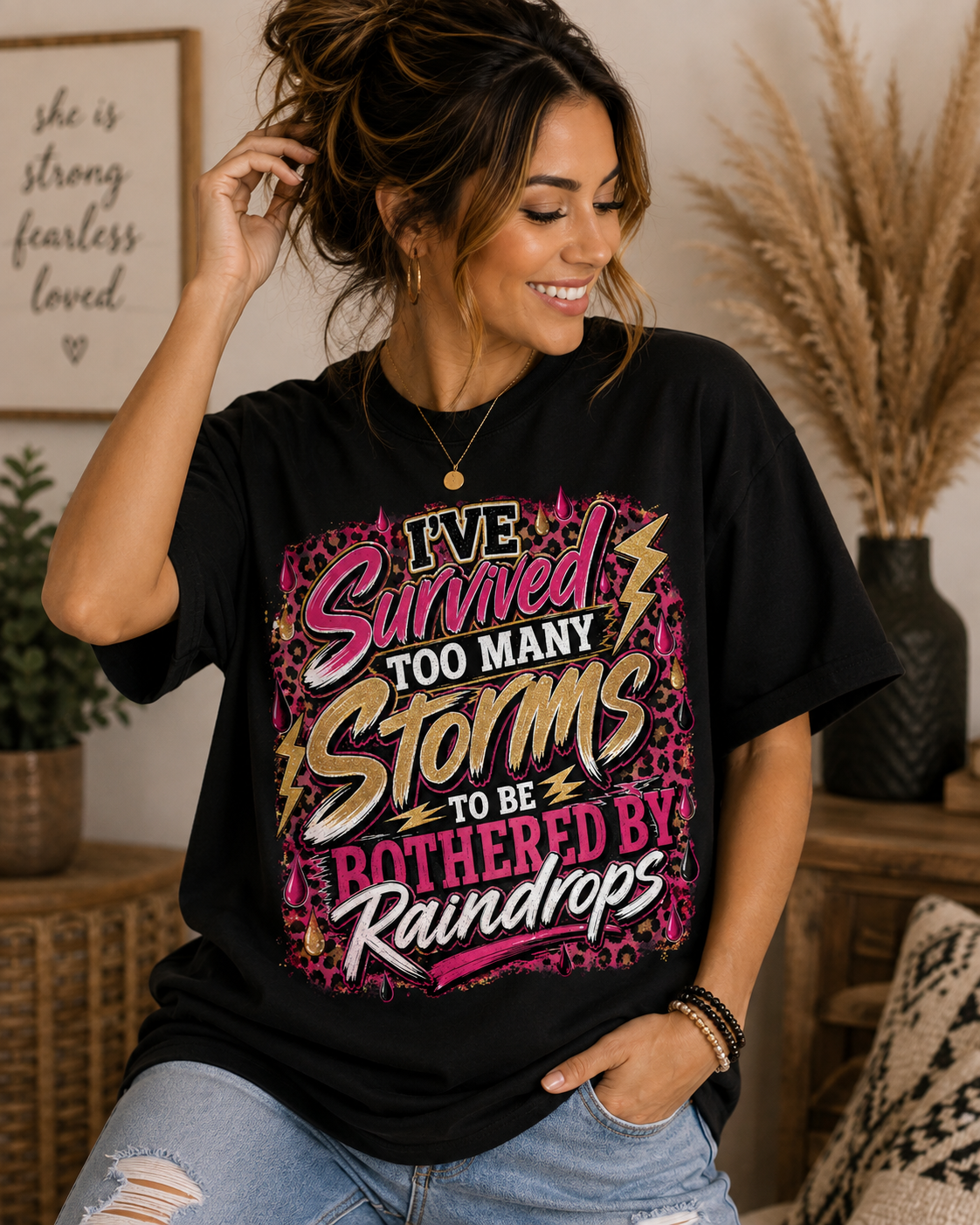 Black: Woman modeling a black I’ve Survived Too Many Storms to Be Bothered by Raindrops graphic T-shirt with pink, gold, leopard-print, lightning, and raindrop details.