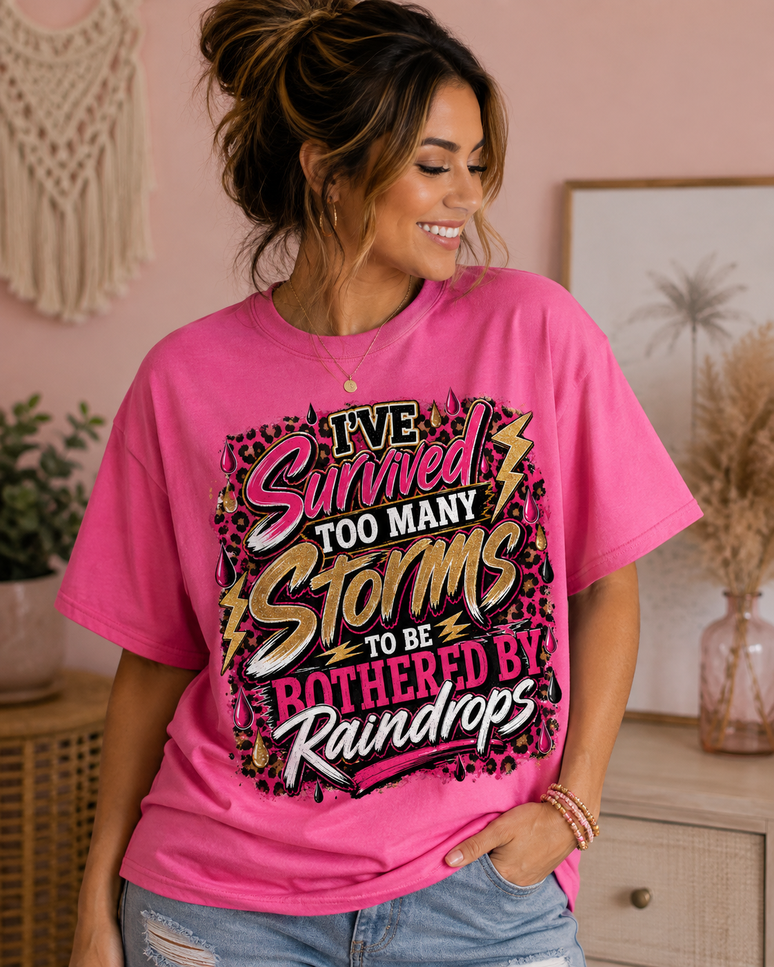 Pink: Woman modeling a hot pink I’ve Survived Too Many Storms to Be Bothered by Raindrops graphic T-shirt with black, white, gold, leopard-print, lightning, and raindrop details.