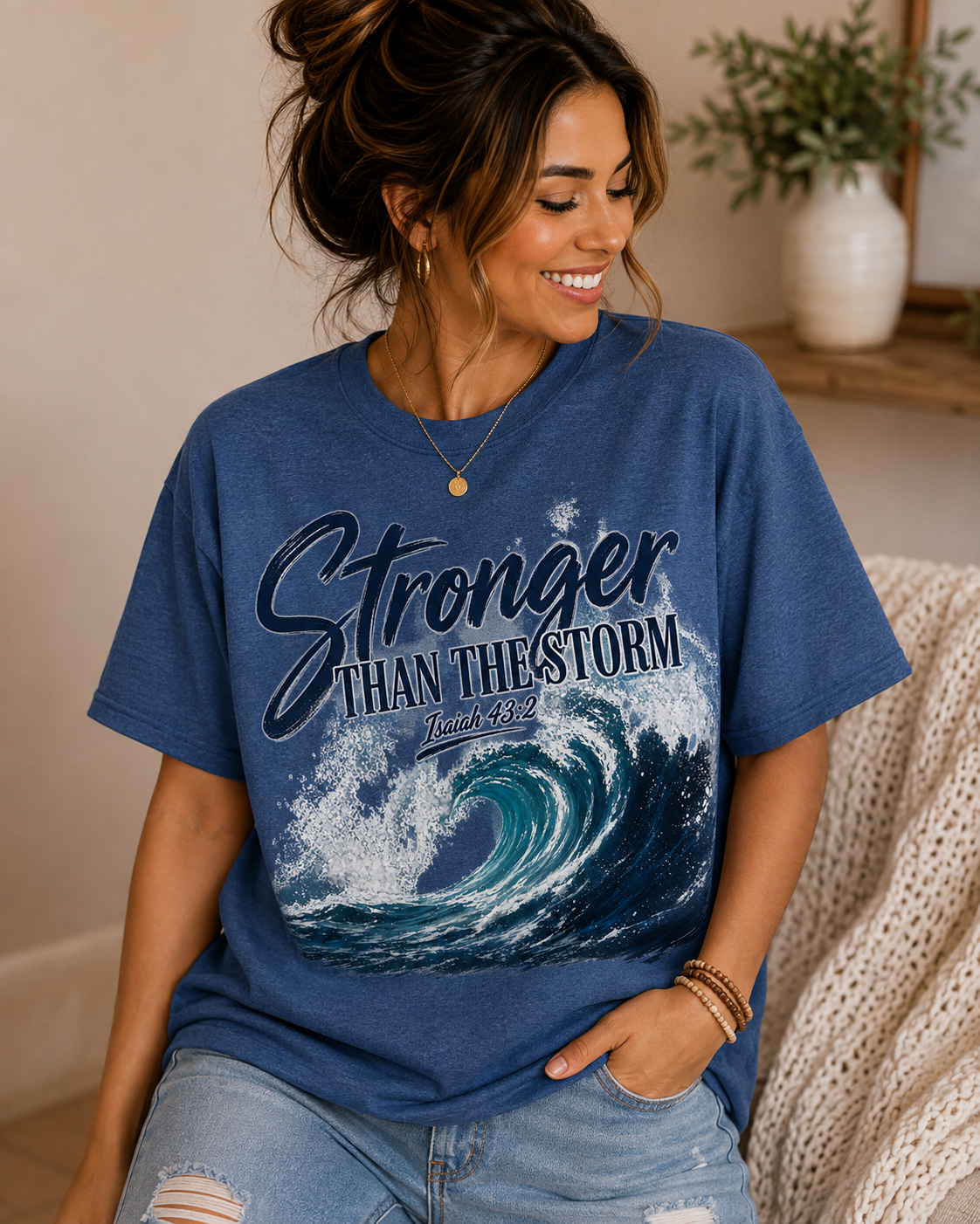 A woman wearing a heathered blue graphic t-shirt with the text 'Stronger THAN THE STORM' and a wave design, paired with light-wash distressed jeans in a cozy indoor setting.