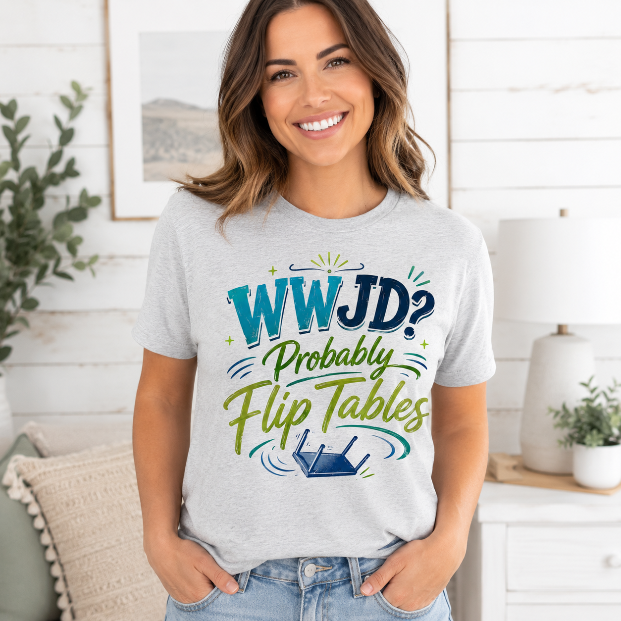 A smiling woman wearing a light gray graphic t-shirt with the text 'WWJD? Probably Flip Tables' in blue and green, paired with light-wash jeans, standing in a bright, modern room with white wood paneling.